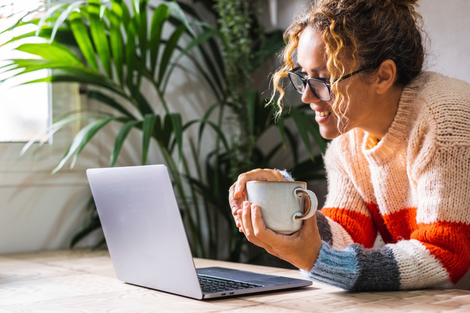 Frau mit Kaffeetasse liest sich Informationen am Laptop durch und lächelt